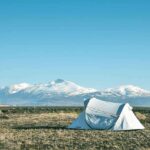 Tente blanche sur une prairie avec des montagnes enneigées en arrière-plan sous un ciel bleu