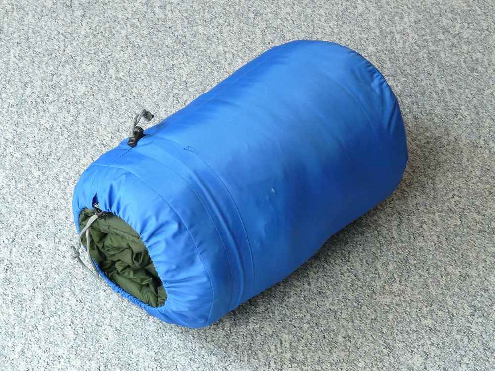 Sac de couchage bleu compressé avec lien de serrage, posé sur sol gris