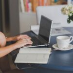 Une personne travaille sur ordinateur à table, avec tasse, cahier et fleurs