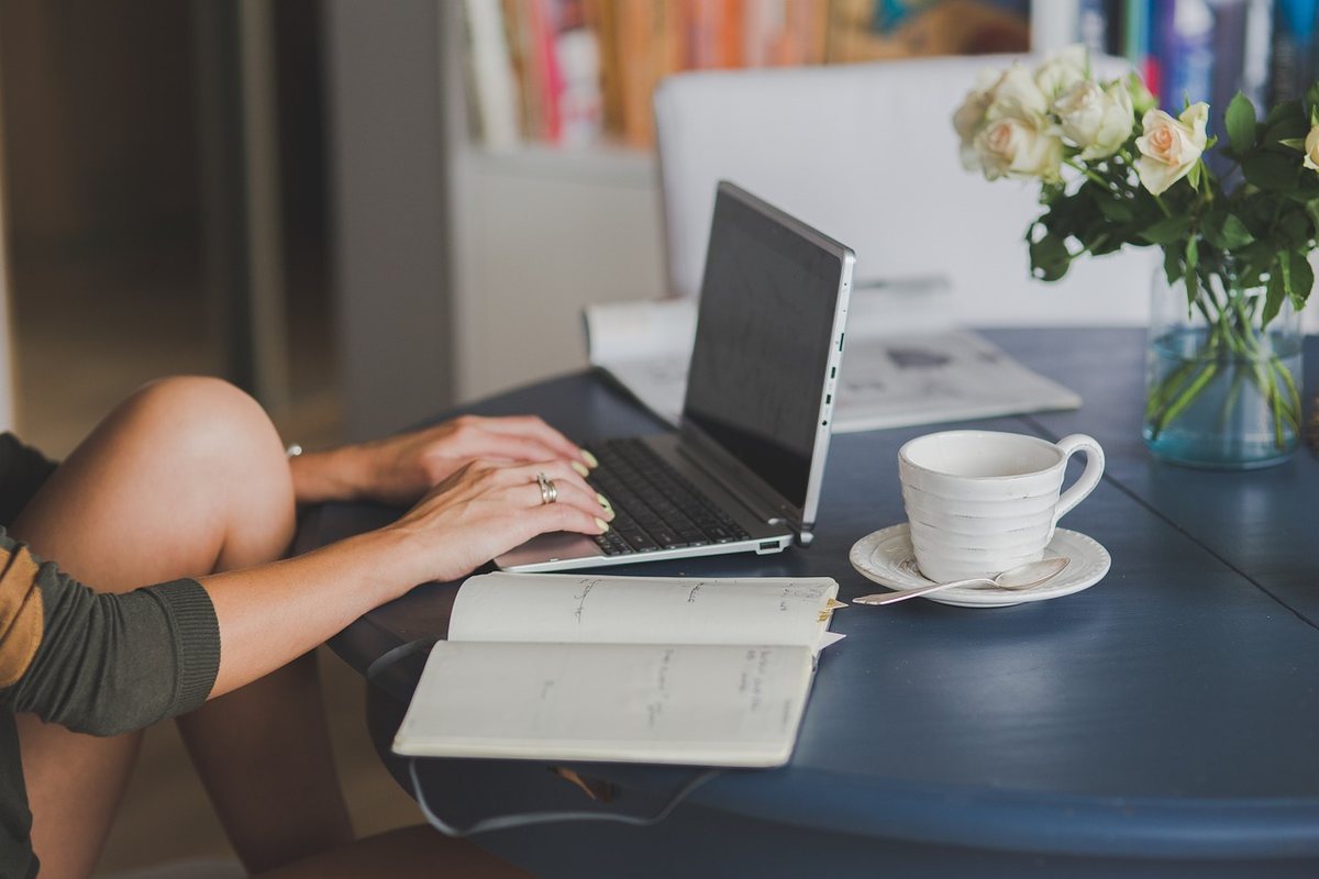 Une personne travaille sur ordinateur à table, avec tasse, cahier et fleurs